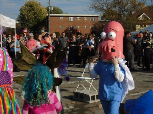 A crowd at a festive event with people in costumes, including a character in a large pink headpiece.
