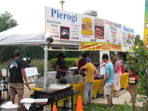 A food stand selling pierogi, sausage, and baked potatoes, with customers ordering and staff preparing food.