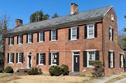 A two-story brick house with black shutters, a front porch, and a well-maintained lawn under a clear blue sky.