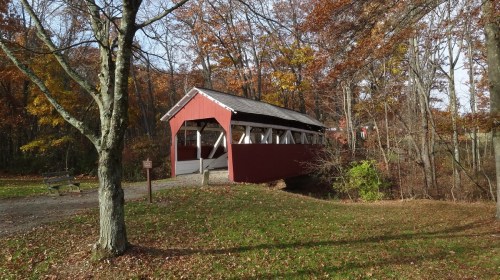 A red covered bridge surrounded by autumn trees and fallen leaves, crossing a small stream in a peaceful setting.