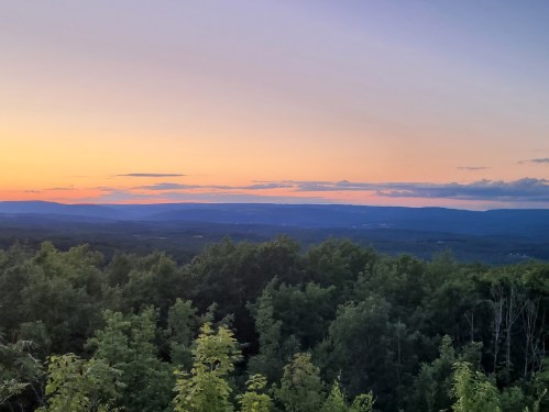 A serene landscape at sunset, featuring rolling hills and a colorful sky above a forested area.