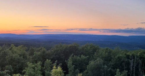 A serene sunset over rolling hills, with vibrant orange and purple hues in the sky and lush green trees in the foreground.
