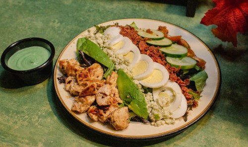 A colorful salad with grilled chicken, hard-boiled eggs, greens, cucumbers, tomatoes, and a side of green dressing.