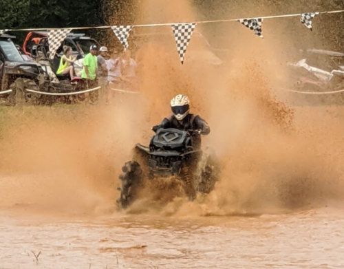 A rider on an ATV splashes through muddy water, sending up a spray of orange-brown dirt, with spectators in the background.