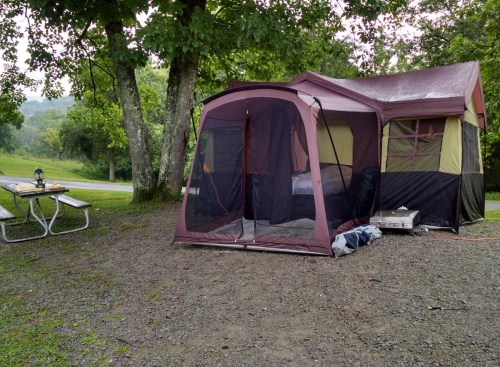 A tent set up in a grassy area, surrounded by trees, with a picnic table nearby and a camping stove visible.