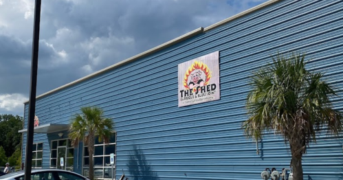 Exterior of a blue building with a sign reading "The Shed" and palm trees in front under a partly cloudy sky.
