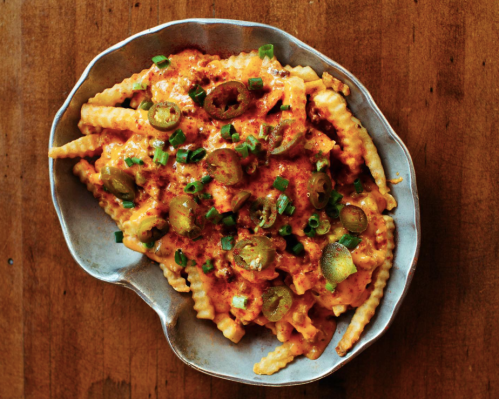 A bowl of crinkle-cut fries topped with cheese sauce, jalapeños, and green onions on a wooden surface.