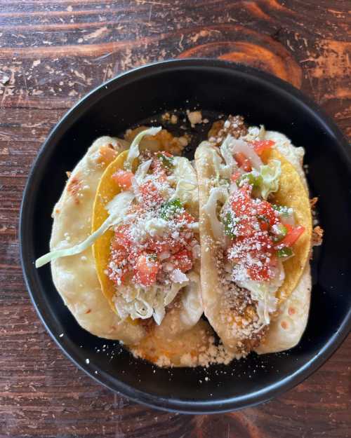 Two tacos on a black plate, filled with lettuce, tomatoes, cheese, and a crispy shell, served on a wooden table.