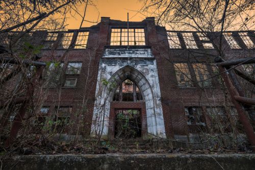 Abandoned brick school building with Gothic architecture, overgrown with plants, under a sunset sky.