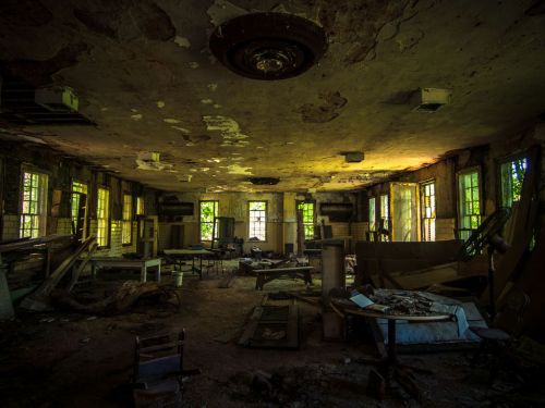 Abandoned room with peeling walls, scattered furniture, and sunlight filtering through dusty windows.