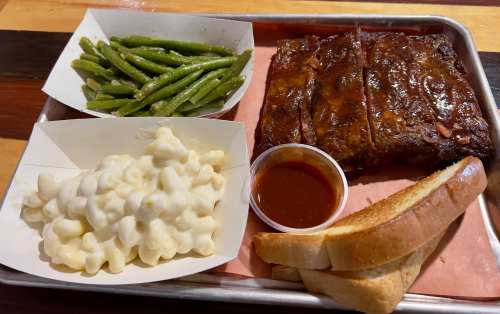 A tray with barbecue ribs, macaroni and cheese, green beans, a side of sauce, and two slices of toasted bread.