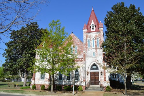 Historic brick church with a tall red roof and steeple, surrounded by green trees and a clear blue sky.