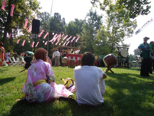 Two people sit on the grass, watching a festival with drums and colorful decorations in a sunny park setting.