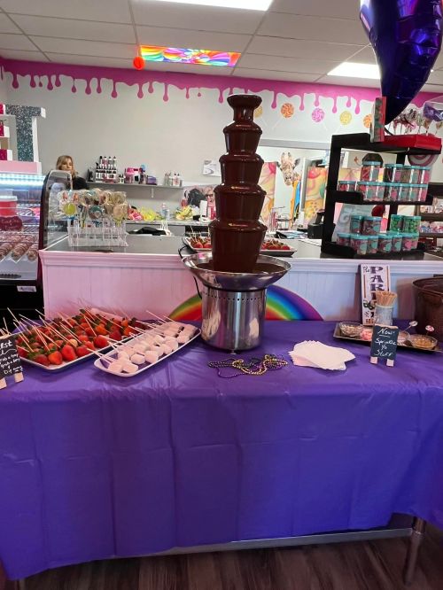 A chocolate fountain surrounded by fruit skewers and treats on a purple tablecloth in a colorful shop.