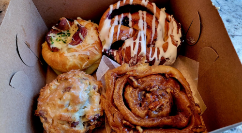 A box containing four baked goods: a cinnamon roll, a pastry with bacon, and two sweet rolls with icing.