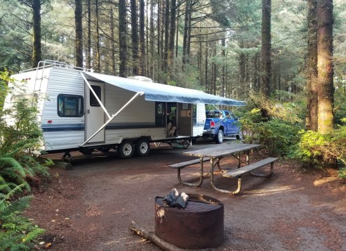 A camper trailer parked in a forested area, with a picnic table and fire pit nearby.