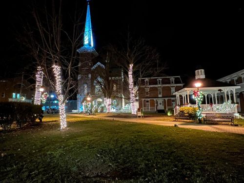 A church and nearby buildings decorated with festive lights at night, surrounded by trees and a grassy area.