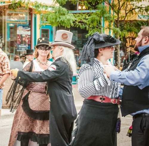A group of people in vintage attire dancing at a lively outdoor event, surrounded by colorful storefronts.