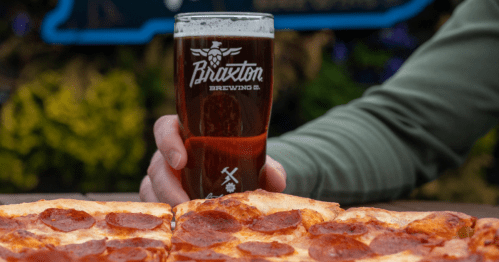 A hand holds a glass of dark beer next to a pepperoni pizza on a wooden table, with greenery in the background.
