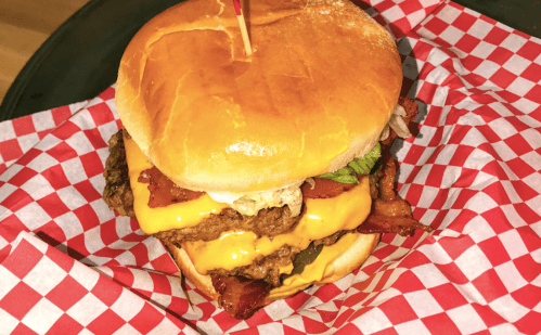 A stacked cheeseburger with bacon, lettuce, and tomato on a toasted bun, served in a red and white checkered paper.