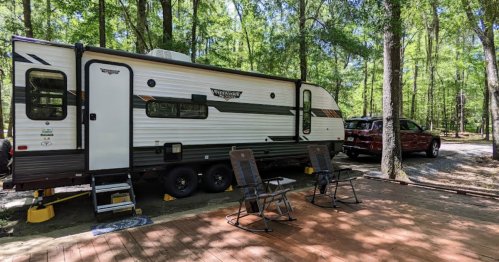 A parked travel trailer in a wooded area, with two chairs and a truck nearby on a wooden deck.