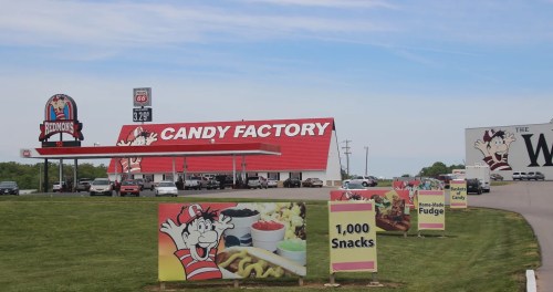 A colorful candy factory with a red roof, featuring signs for snacks and fudge, located along a highway.
