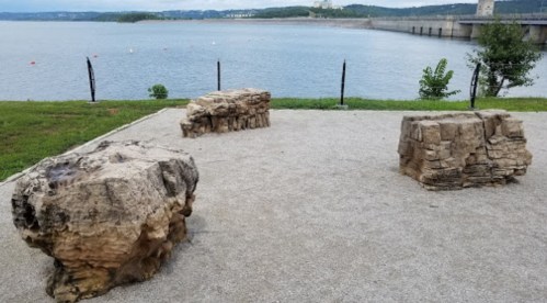 Two large stone benches on a gravel area by a calm lake, with greenery and a distant dam visible.