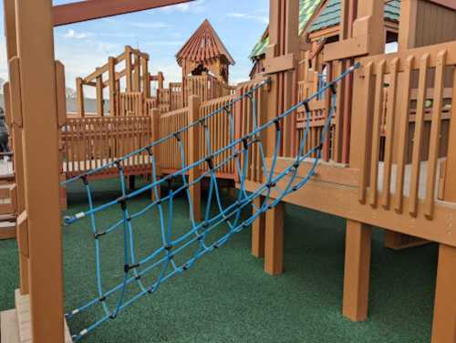 A playground structure featuring wooden platforms and a blue rope bridge connecting them, set on green turf.