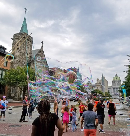A group of people enjoying large soap bubbles near a church and a government building under a cloudy sky.