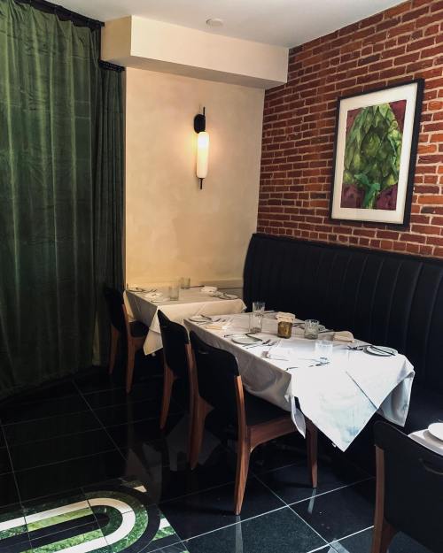 A cozy restaurant corner with a black leather booth, white tablecloths, and a brick wall adorned with artwork.