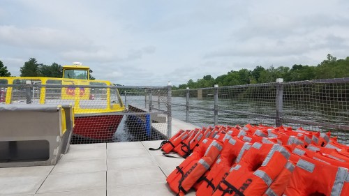 A yellow boat docked by a river, with a pile of orange life jackets in the foreground and trees in the background.