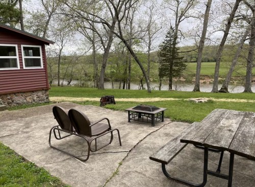 A cozy outdoor space with two chairs, a fire pit, and a picnic table, overlooking a river and lush greenery.