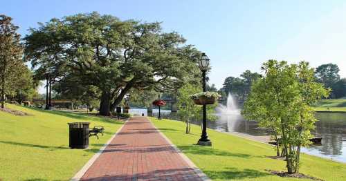 A scenic park pathway lined with trees, benches, and a fountain beside a calm waterway on a sunny day.