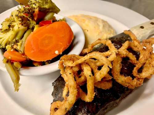 A plate featuring grilled steak topped with crispy onions, served with mixed vegetables and a biscuit.