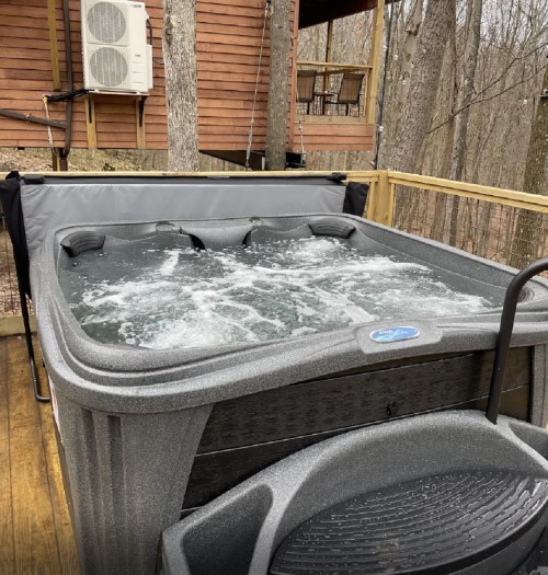 A hot tub with bubbling water on a wooden deck surrounded by trees and a cabin in the background.