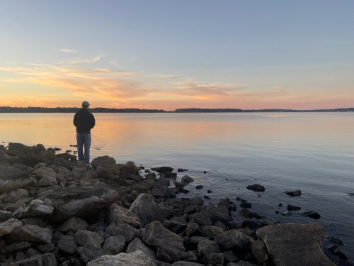 A person stands on rocky shore, fishing at sunset, with calm water and colorful sky in the background.