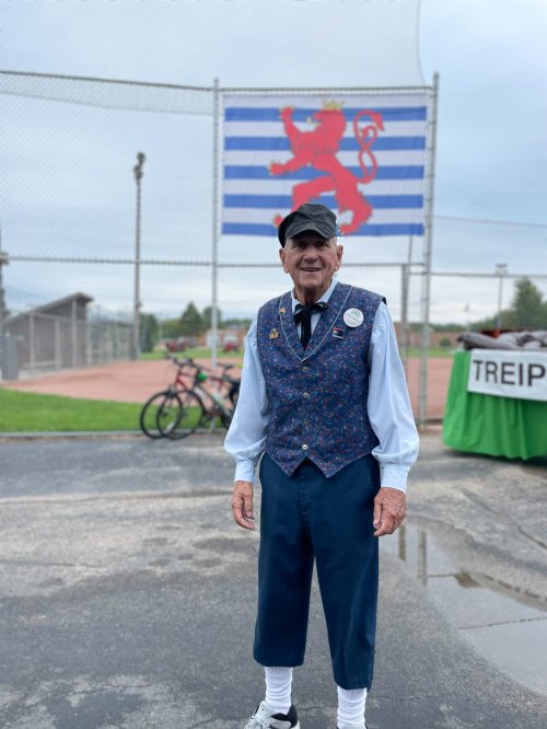 An elderly man in a patterned vest and cap stands in front of a flag at a sports field, with bicycles in the background.