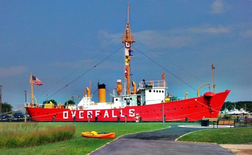 A red lighthouse ship named "Overfalls" docked near a grassy area, with a kayak in the foreground and a clear blue sky.