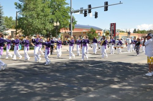 A marching band in purple uniforms parades down a street, with spectators watching from the sidewalk.