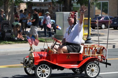 A man in a fez waves from a small red parade car decorated with American flags. Spectators watch in the background.