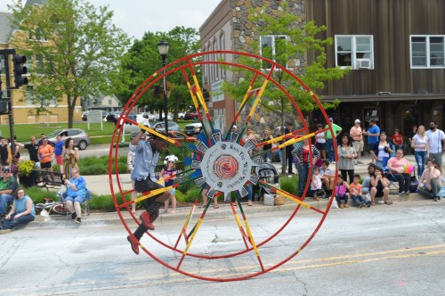 A performer in a large wheel entertains a crowd during a parade, with onlookers watching from the sidewalk.
