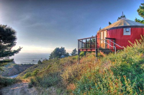 A red yurt on a hillside overlooking the ocean, surrounded by greenery and trees under a cloudy sky.