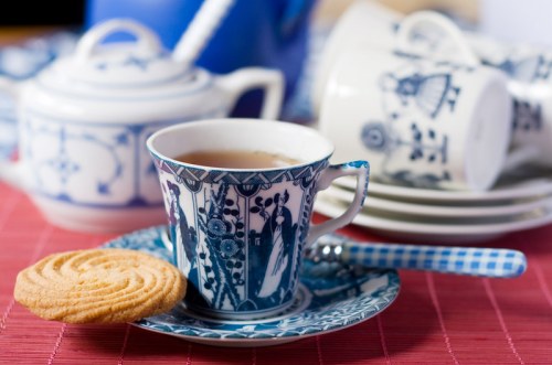 A blue and white patterned teacup with tea, a cookie on the saucer, and a teapot in the background.