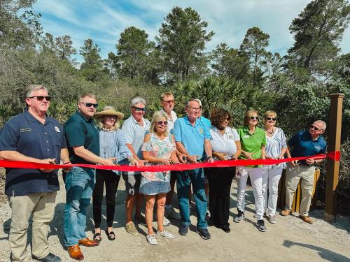 A group of people stands together, holding a red ribbon for a ceremonial ribbon-cutting event in a natural setting.