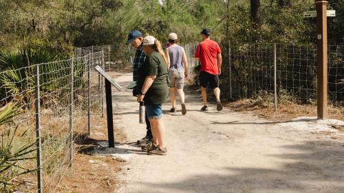 A group of people walking on a dirt path, with one person reading a sign near a fence in a wooded area.