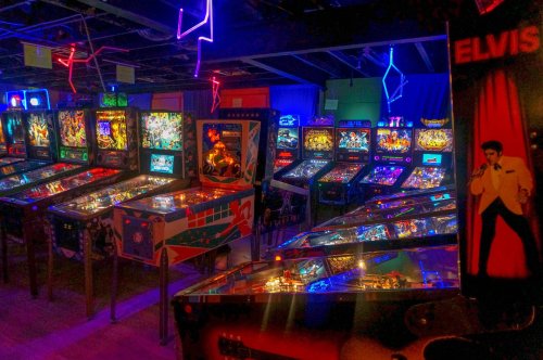 A vibrant arcade filled with colorful pinball machines and neon lights, featuring an Elvis-themed game on the right.