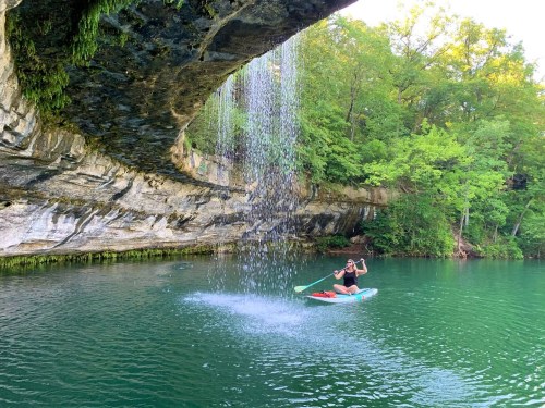 A person kayaking under a rocky overhang with a waterfall, surrounded by lush green trees and calm water.