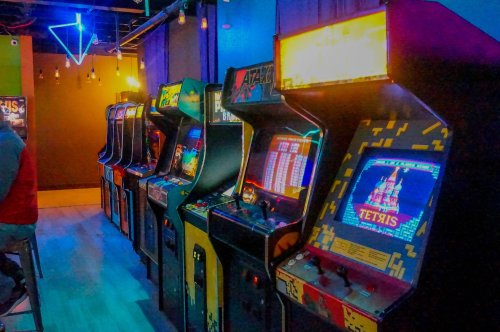 A row of vintage arcade machines in a colorful gaming space, featuring titles like Tetris and Atari.