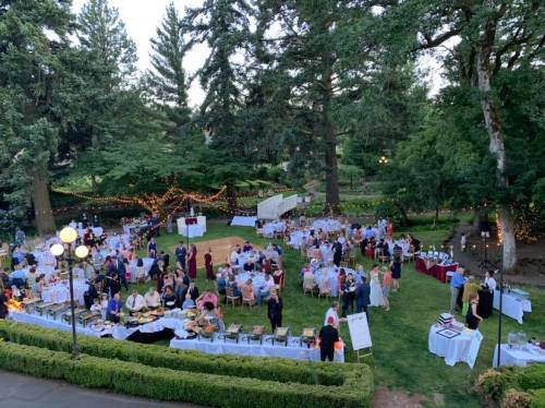 A lively outdoor event with guests dining at tables surrounded by trees and string lights, set in a garden.
