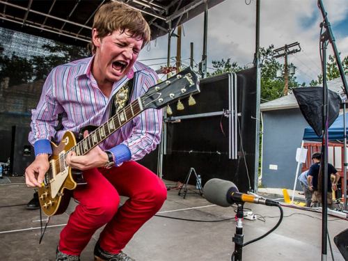 A guitarist passionately performs on stage, wearing a striped shirt and red pants, with a microphone nearby.